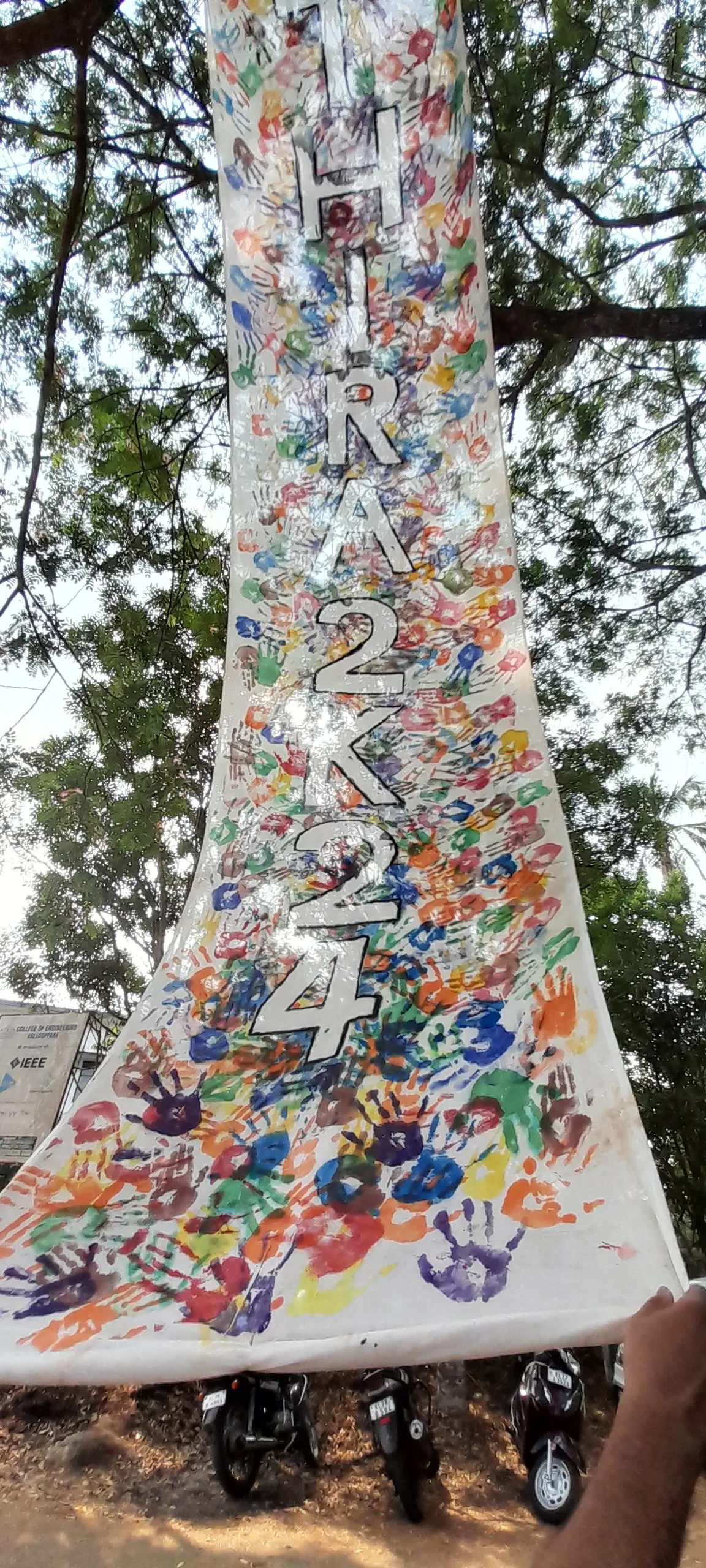 A large cotton cloth with height hung from a tree. There are a lot of handprints on the cloth with different cloth. Thira 2k24, name of the arts fest is written on the cloth on top of the handprints.