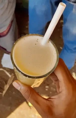 A hand holding a glass filled to the brim with cold sugarcane juice made from sugarcane, lemon juice,ginger and sugar. There is also a paper straw in the glass.